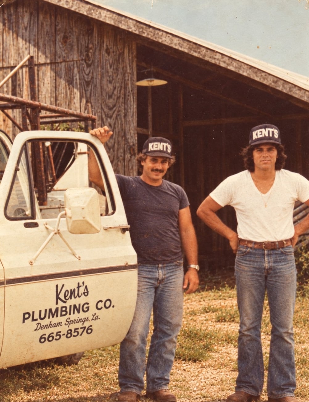 Jimmy Joe and Woody Kent in Kent’s caps standing beside the original Kent’s Plumbing Co. truck outside a wooden barn in Denham Springs, Louisiana